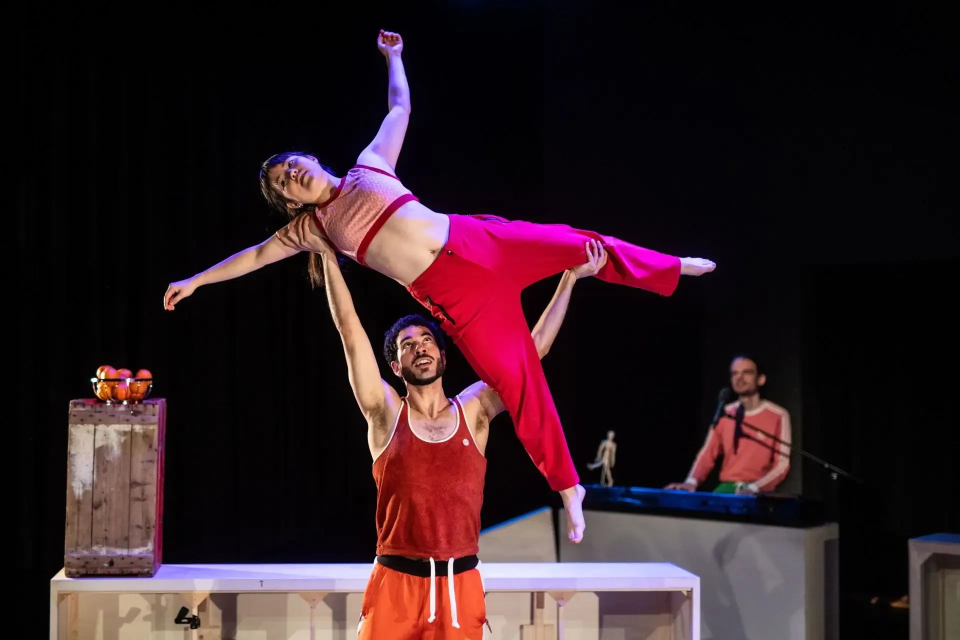 Een danser houdt een andere danser hoog in de lucht. Ze hebben beide felrode kleren aan.